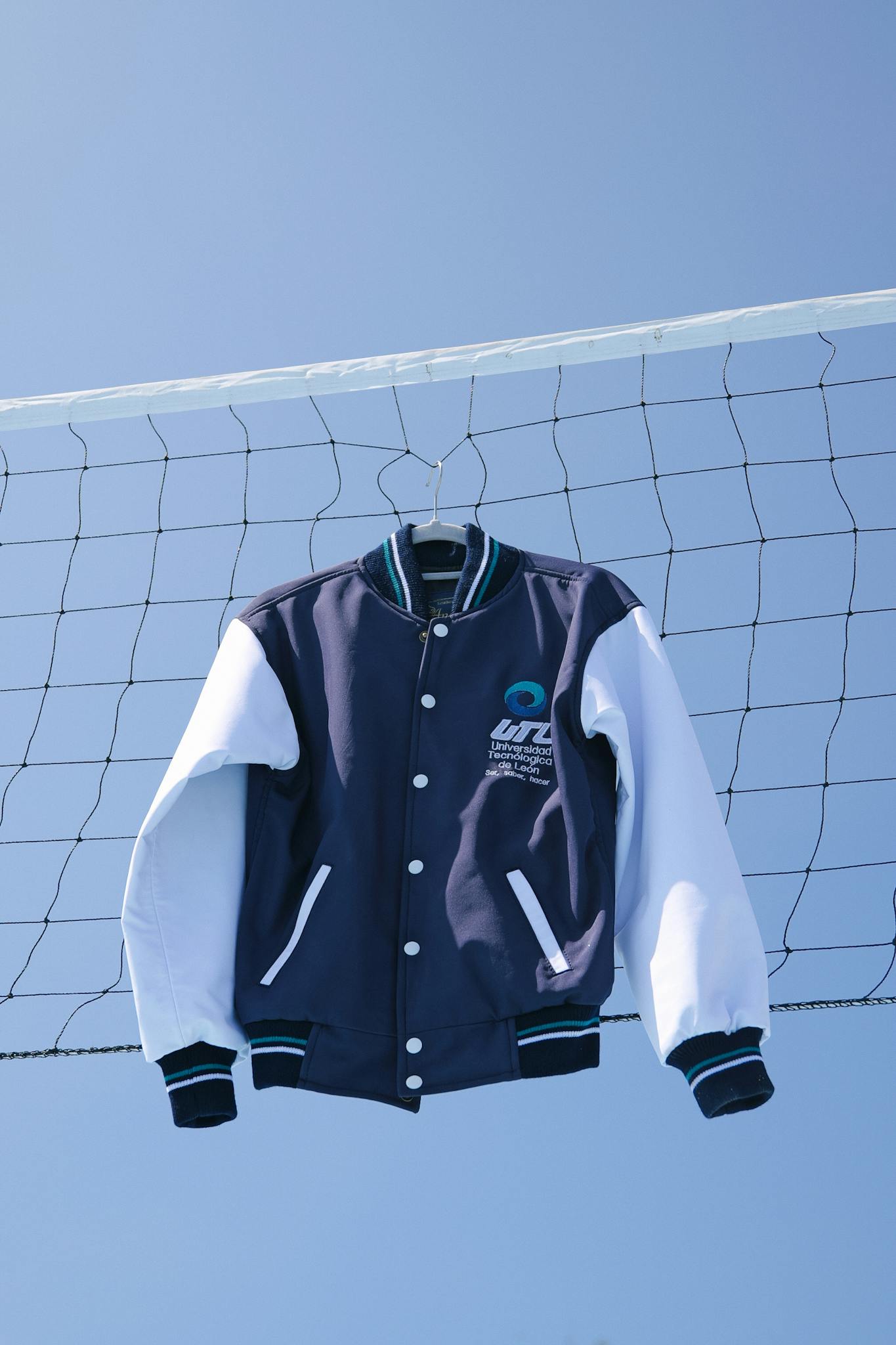 A classic varsity jacket hanging on a volleyball net against a clear blue sky in León, México.