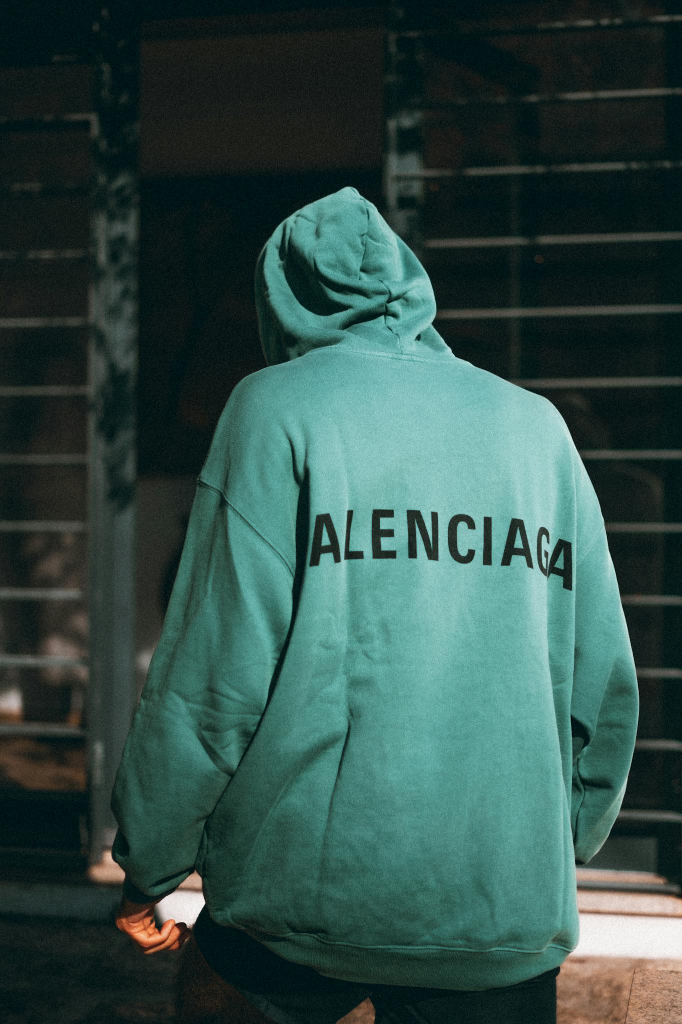 Model in green hoodie with logo stands in front of grid gate.