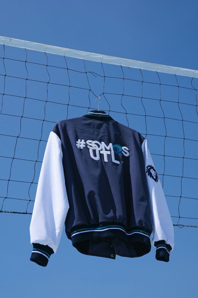 Stylish blue varsity jacket hanging on a sports net against a clear sky.