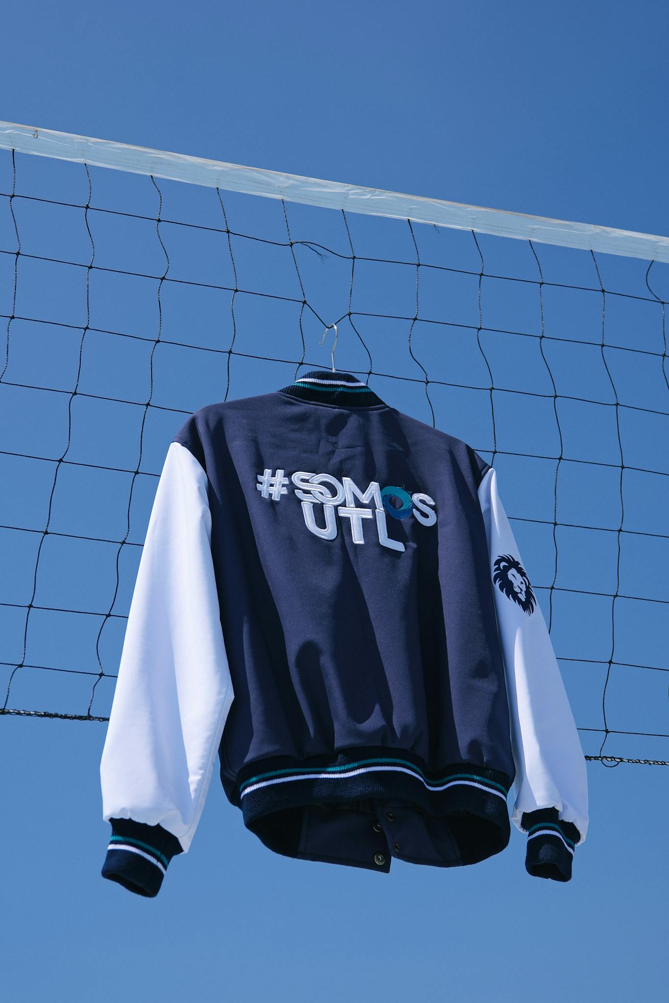 Stylish blue varsity jacket hanging on a sports net against a clear sky.